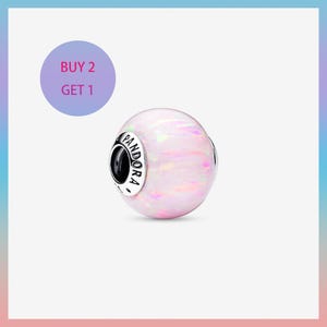 Op de afbeelding: Een ronde, opalescerende bedel met parelmoer roze en witte tinten. De bedel heeft het woord "PANDORA" in zwarte letters op een zilveren band. Een paarse cirkel met de tekst "KOOP 2 KRIJG 1" staat linksboven.
