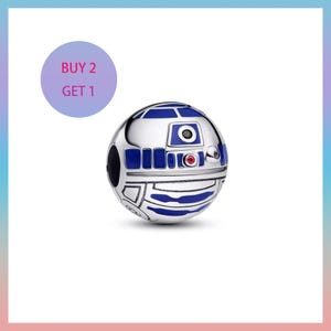 May include: A silver charm depicting the Star Wars droid R2-D2, with blue and black detailing. The spherical charm has a red accent. A purple circle in the upper left corner displays the text "BUY 2 GET 1".