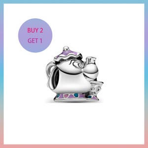 Puede incluir: Un charm plateado con forma de tetera con tapa y base moradas, una taza pequeña y una botella. El charm tiene un diseño de dibujos animados. El texto "BUY 2 GET 1" está en un círculo morado en la esquina superior izquierda.