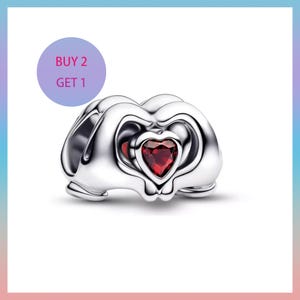 Puede incluir: Un charm de color plateado con un coraz&oacute;n rojo en el centro. El charm tiene un dise&ntilde;o &uacute;nico con elementos curvos y calados. Un c&iacute;rculo p&uacute;rpura con el texto "BUY 2 GET 1" est&aacute; en la esquina superior izquierda.