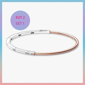 Puede incluir: Una pulsera de dos tonos con un cierre plateado y una banda de oro rosa. La parte plateada tiene grabado el nombre de la marca. La imagen incluye un círculo morado con el texto "BUY 2 GET 1".