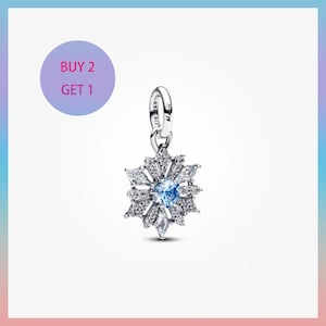 May include: A silver snowflake charm with a central blue gemstone and clear crystal accents. The charm is suspended from a silver loop. The image includes a purple circle with the text "BUY 2 GET 1".