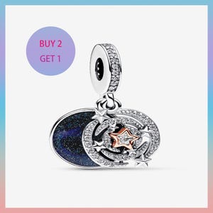 Puede incluir: Un charm de plata con temática celestial. Presenta un óvalo azul oscuro con purpurina, un anillo de piedras transparentes y una estrella de oro rosa. El charm tiene un lazo en la parte superior. El texto "BUY 2 GET 1" está en un círculo morado.