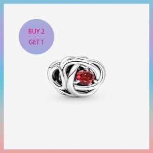 Puede incluir: Un charm de plata con un diseño entrelazado, con una gema roja vibrante en el centro. El fondo presenta un círculo morado con el texto "BUY 2 GET 1".
