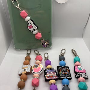 Puede incluir: Una colección de colgantes de teléfono con cuentas con varios dijes. Un dije presenta un fantasma leyendo un libro. Otros dijes incluyen una taza de café, un libro y frases como "Always Freakin Busy Reading Trash".