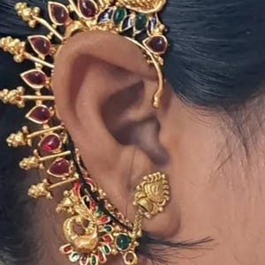 May include: Ornate gold ear cuff with red, green, and blue accents. The jewelry features a peacock design and is adorned with small red gemstones. A dangling earring with a gold ball and white beads is also visible.