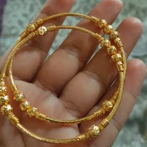 Puede incluir: Dos pulseras doradas con detalles intrincados. Las pulseras presentan un diseño de cuerda trenzada y están adornadas con pequeños acentos redondos y con cuentas. El color dorado es brillante y reflectante, lo que sugiere un acabado pulido. Las pulseras se muestran sobre un fondo neutro.