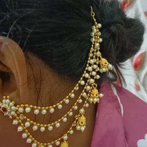 May include: Gold-toned hair jewelry with multiple strands of small, white pearls. The piece features ornate gold pendants and a small red accent. The jewelry is designed to be worn on the side of the head, with a hook for attachment.