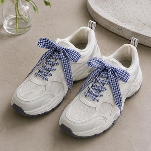 May include: A pair of white athletic sneakers featuring mesh and synthetic leather panels, thick rubber soles, and blue and white gingham patterned fabric laces tied in bows at the top of each shoe, with a small striped pull tab at the heel.