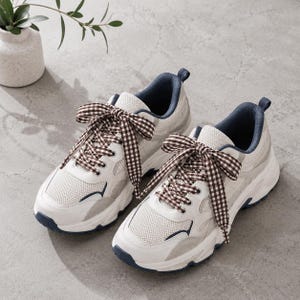 May include: A pair of white and light gray sneakers featuring mesh panels, dark blue interior lining, and brown and white gingham patterned fabric laces tied in large bows. The sneakers have thick white rubber soles with dark blue accents.