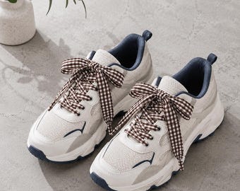 May include: A pair of white and light gray sneakers featuring mesh panels, dark blue interior lining, and brown and white gingham patterned fabric laces tied in large bows. The sneakers have thick white rubber soles with dark blue accents.