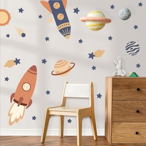 Peut inclure: Décoration de chambre d'enfant sur le thème de l'espace, avec des fusées, des planètes et des étoiles sur un mur gris clair. Une chaise et une commode en bois sont au premier plan, avec un lapin en peluche et un bloc géométrique.