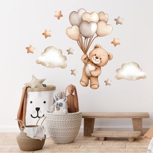 Puede incluir: Un conjunto de calcomanías de pared para guardería con un oso de peluche marrón sosteniendo globos en forma de corazón. El conjunto incluye nubes, estrellas y un contenedor de almacenamiento con temática de oso. Un banco de madera y cestas llenas de peluches completan la escena.