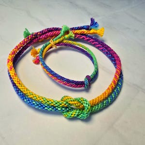 May include: A set of two braided cord accessories, one necklace and one bracelet, featuring a vibrant multi-colored pattern of yellow, green, blue, purple, and pink threads. Each piece is tied with a decorative knot and has loose frayed ends.