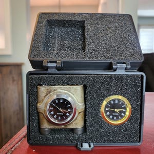 May include: An open black case with a foam interior holds two quartz clocks. One clock is gold-toned with a black face and Roman numerals. The other clock is a smaller, round gold-toned clock with a black face.