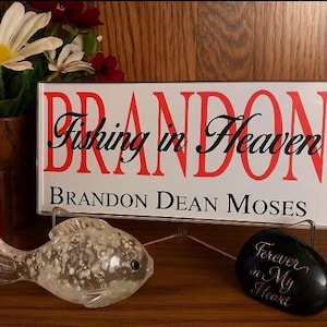 May include: A memorial sign with the name "Brandon" in red, and the words "Fishing in Heaven" in black script. A clear glass fish and a black stone with "Forever in My Heart" are in front of the sign.
