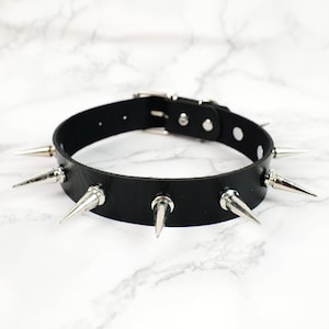Op de afbeelding: Een zwarte leren choker ketting met zilverkleurige metalen spikes. De choker heeft een gespsluiting en staat tegen een witte marmeren achtergrond. De spikes zijn gelijkmatig verdeeld.
