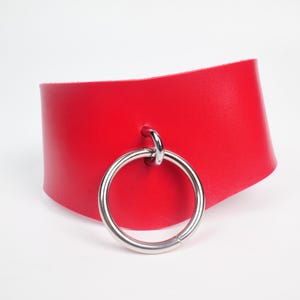 Könnte beinhalten: Ein leuchtend rotes Lederhalsband mit einem großen, silberfarbenen Metallring. Das Halsband ist ein breites Band, das um den Hals getragen wird. Der Ring ist vorne befestigt und verleiht einen kühnen, kantigen Look. Der Hintergrund ist weiß.