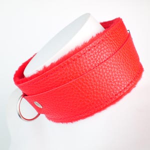 May include: A red leather collar with a soft, plush red interior. The collar has a silver metal ring and a buckle closure. The collar is shown on a white form.