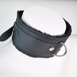 May include: A black leather collar with a soft, black fur lining. The collar has a silver-colored metal ring and a strap with a snap closure. The collar is designed to be worn around the neck.