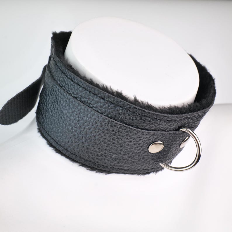 May include: A black leather collar with a soft, black fur lining. The collar has a silver-colored metal ring and a strap with a snap closure. The collar is designed to be worn around the neck.