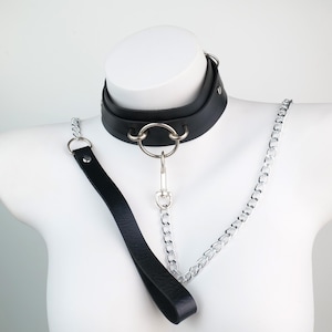 Op de afbeelding: Zwarte leren halsband met een zilveren metalen ring en kettingriem. De halsband is dik en heeft een bijpassende zwarte leren riem. De ketting is zilverkleurig en wordt met een metalen sluiting aan de halsband bevestigd.