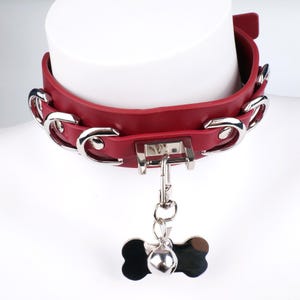 May include: A red leather collar with silver metal detailing and a bone-shaped tag. The collar has a buckle fastening and decorative chain links. A small silver bell hangs from the tag.