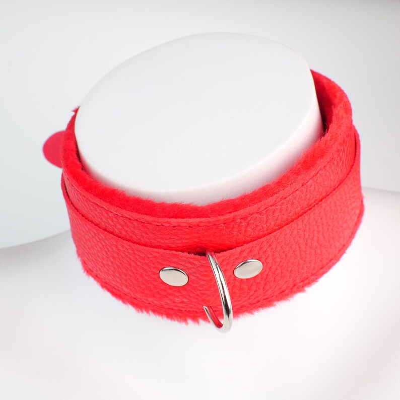 May include: A red leather collar with a soft, plush interior. The collar features a silver metal ring and two silver rivets. The collar is displayed on a white mannequin head.