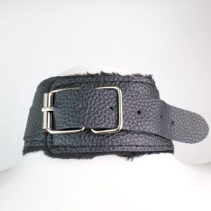 May include: Black leather choker with a textured finish and a silver-colored buckle. The choker has a belt-style closure and a soft, furry lining. The design is simple and elegant.