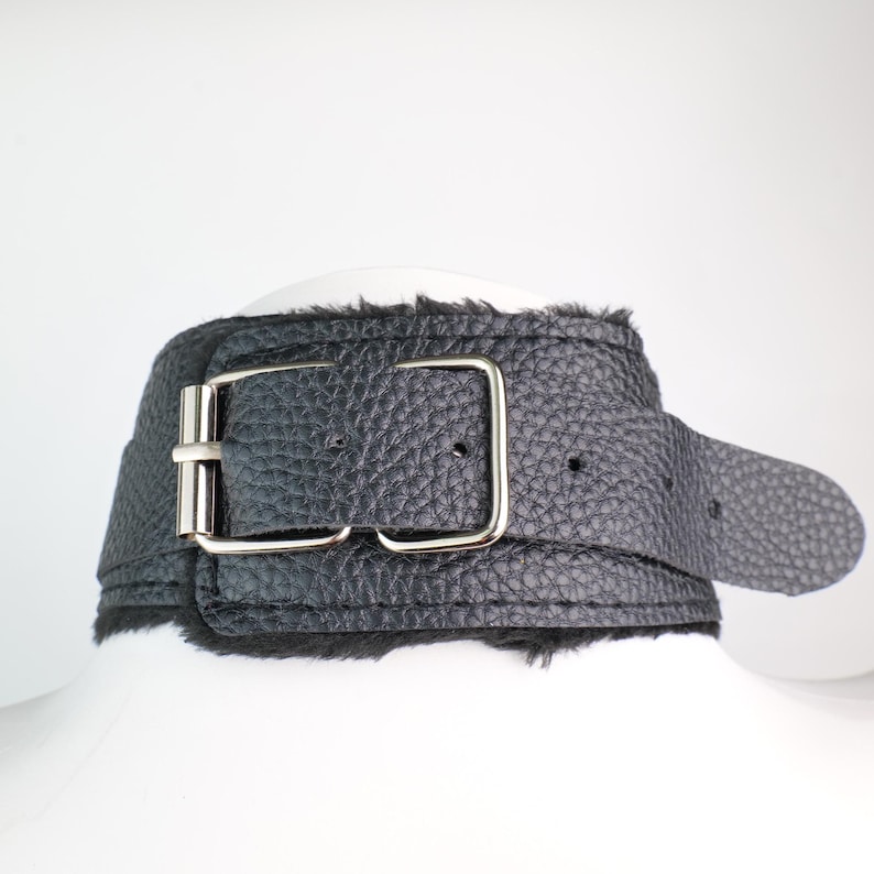 May include: Black leather choker with a textured finish and a silver-colored buckle. The choker has a belt-style closure and a soft, furry lining. The design is simple and elegant.