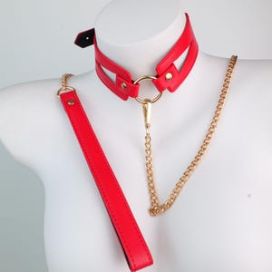 May include: A red leather collar with a gold-colored chain and a matching leash. The collar features a geometric design with gold-colored hardware. The leash is a long, rectangular strap.