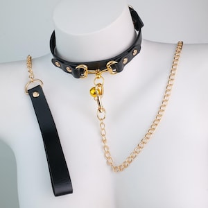 May include: A black leather collar with gold-colored hardware, including a chain and a small bell. The collar is displayed on a white mannequin. A matching black leather leash is also included.