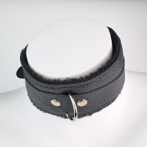 May include: Black leather collar with a soft, plush interior lining. The collar features a silver-colored metal ring and two silver-colored metal studs. The collar is designed to be worn around the neck.