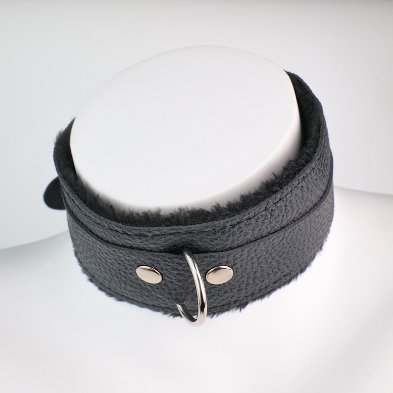 May include: Black leather collar with a soft, plush interior lining. The collar features a silver-colored metal ring and two silver-colored metal studs. The collar is designed to be worn around the neck.