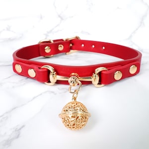 May include: Red leather collar with gold-colored hardware and a decorative bell. The collar features a buckle closure and gold-colored rivets. The bell is a filigree design. This collar is designed for pets.