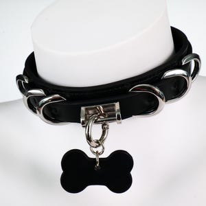 May include: Black leather collar with silver-tone metal accents and a bone-shaped tag. The collar features a buckle closure and large, curved metal links. The tag hangs from a metal ring.