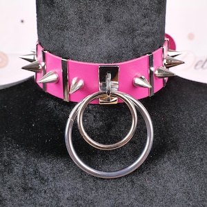 May include: A hot pink leather choker necklace with silver-colored metal spikes and two large silver rings. The choker is displayed on a black mannequin head.