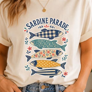Könnte beinhalten: Ein cremefarbenes Kurzarm-T-Shirt mit einem Grafikdruck von fünf übereinander gestapelten Fischen mit verschiedenen Mustern wie Vichy-Karo, Wellen, Blumen, Punkten und Streifen. Der Text "SARDINE PARADE" ist in blauen Buchstaben darüber gebogen.