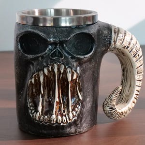 May include: A novelty mug with a skull design. The mug is dark gray with a stainless steel rim and a handle shaped like a horn. The skull features large eye sockets and a wide, toothy grin.