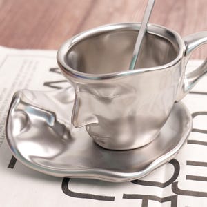 Può includere: Un set di tazza e piattino da caffè color argento con un design a volto unico. La tazza ha un manico e un cucchiaino all'interno. Il piattino ha un design a volto abbinato. Il set è in metallo e poggia su un giornale.
