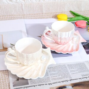 May include: Two ceramic teacups and saucers. The cups and saucers are pearlescent white and pink, shaped like seashells. Each cup has a handle with a pearl accent. The saucers have a pearl accent. The cups are on a newspaper.