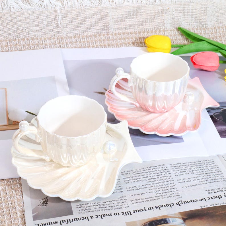 May include: Two ceramic teacups and saucers. The cups and saucers are pearlescent white and pink, shaped like seashells. Each cup has a handle with a pearl accent. The saucers have a pearl accent. The cups are on a newspaper.