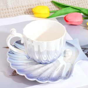 May include: A pearlescent white teacup and saucer set. The cup has a wavy design and a pearl accent on the handle. The saucer is shell-shaped with a pearl accent. The set has a gradient of blue and white.