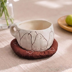 Peut inclure: Une tasse en céramique couleur crème avec un motif de poires dessiné à la main en noir et une petite anse circulaire, posée sur un dessous de verre rond en tissu marron foncé orné d'un motif floral brodé en blanc.