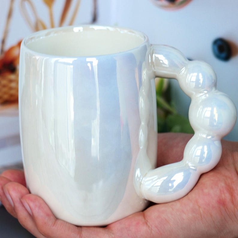 May include: A pearlescent white ceramic mug with a unique handle design. The handle is made of connected, rounded shapes. The mug has a glossy finish and reflects light, creating a shimmering effect. The mug is held in a person's hand.