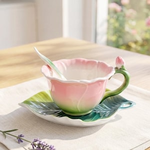 May include: A ceramic teacup and saucer set shaped like a blooming flower. The cup features a pink and green gradient with a green handle topped by a small pink bud. The saucer is shaped like a large green leaf. A matching spoon is inside the cup.