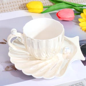May include: A pearlescent white teacup and saucer set. The cup has a scalloped rim and a pearl accent on the handle. The saucer is shaped like a seashell, with a pearl accent. The set is on a white and gray surface with colorful flowers.