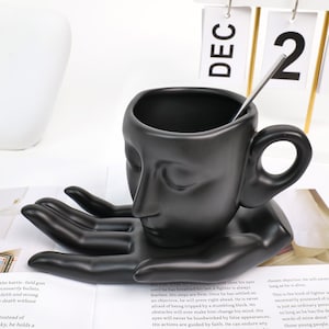 Pode incluir: Uma caneca de cerâmica preta com um design de rosto, sustentada por uma base em forma de mão. Uma colher de prata está dentro da caneca. O calendário mostra 2 de dezembro. Um item decorativo único.