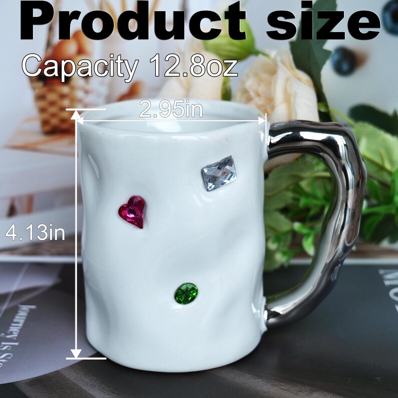 May include: White ceramic mug with a silver handle, adorned with jewel-like embellishments. The mug is 4.13 inches tall, 2.95 inches wide, and has a capacity of 12.8oz. The mug has a textured surface and is decorated with a pink heart, a green circle, and a clear square.