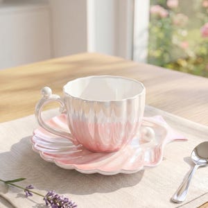 May include: A ceramic teacup and saucer set featuring a scalloped shell design with a pearlescent pink and white finish. The cup has a curved handle with a small sphere at the top, and the saucer includes a decorative white pearl accent.